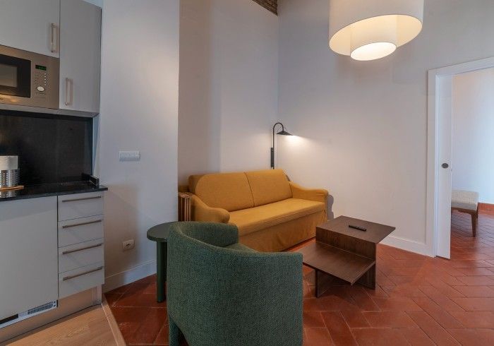 Sala de estar moderna con sofá color mostaza, silla verde, mesa de centro, suelo de terracota y rincón de cocina.