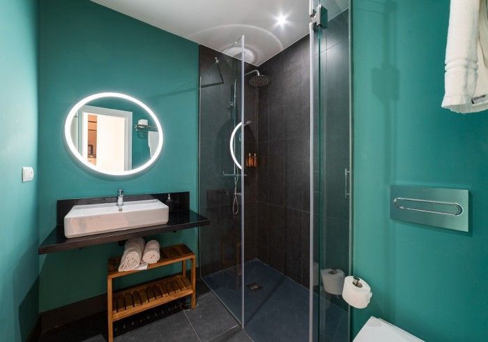 Baño moderno con paredes color turquesa, espejo redondo, lavabo con tocador y cabina de ducha de cristal.