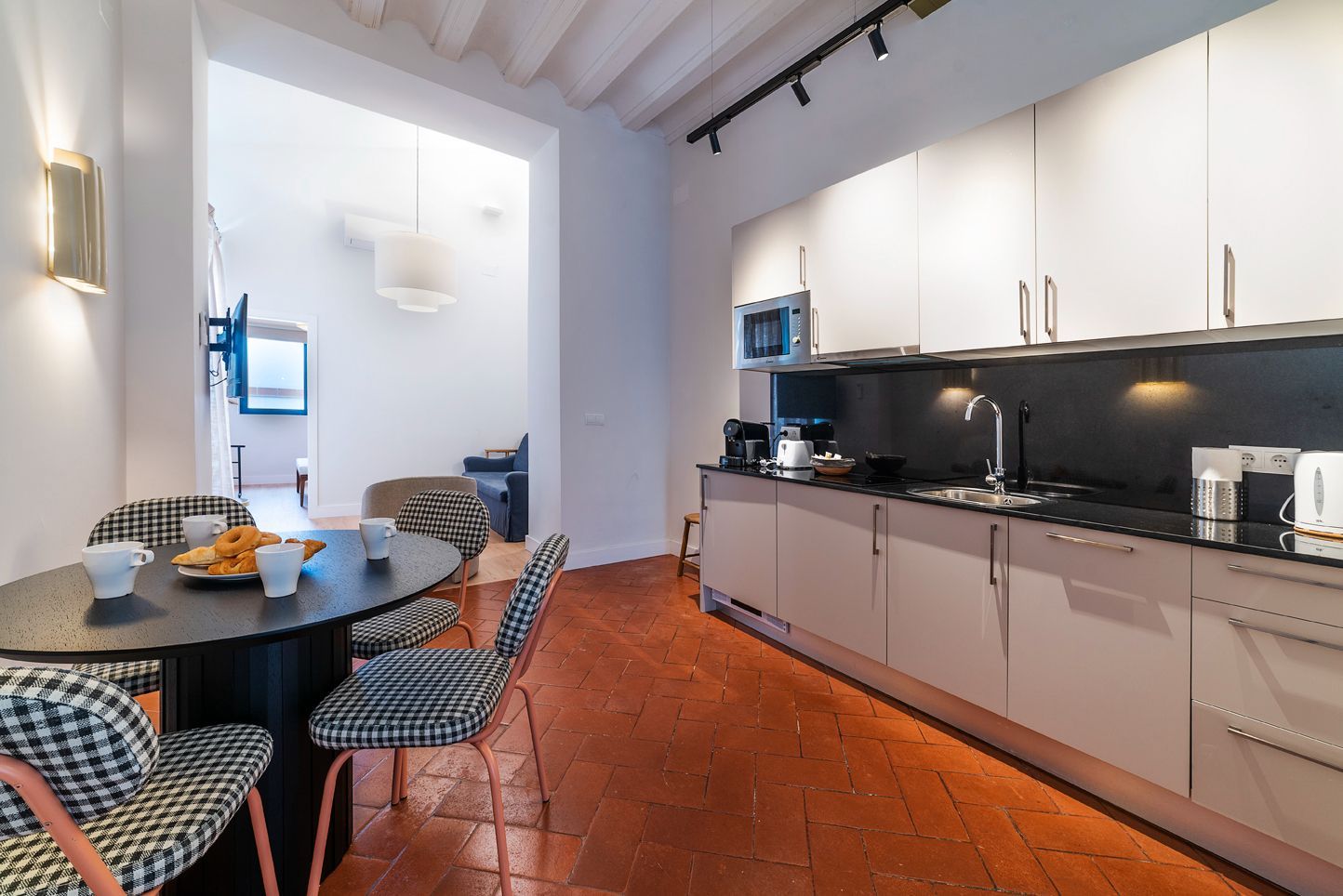 Cocina moderna con mesa de comedor, encimeras negras, armarios blancos y suelo de baldosas de terracota.