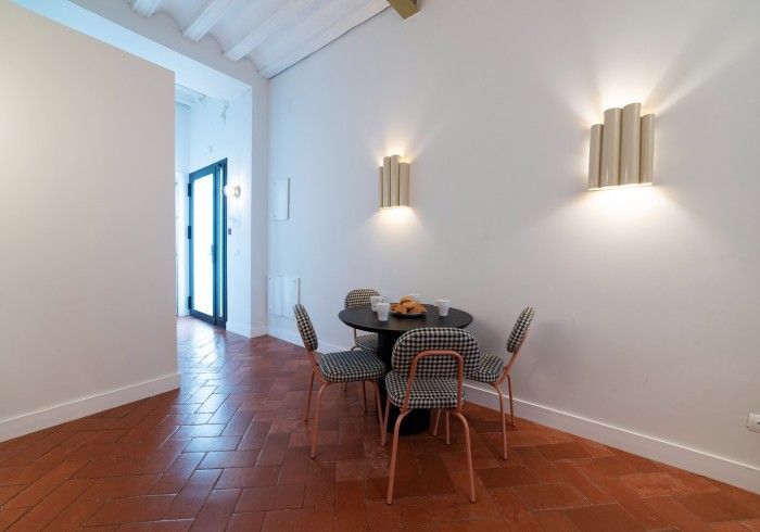 Comedor luminoso con suelo de baldosas de terracota, mesa redonda, sillas y apliques de pared en una habitación blanca.