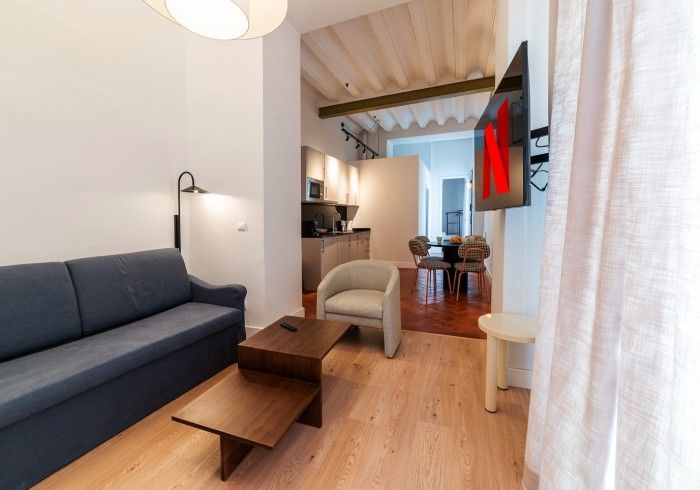 Luminoso salón moderno con sofá azul, mesa de centro de madera, silla beige y televisor montado en la pared.