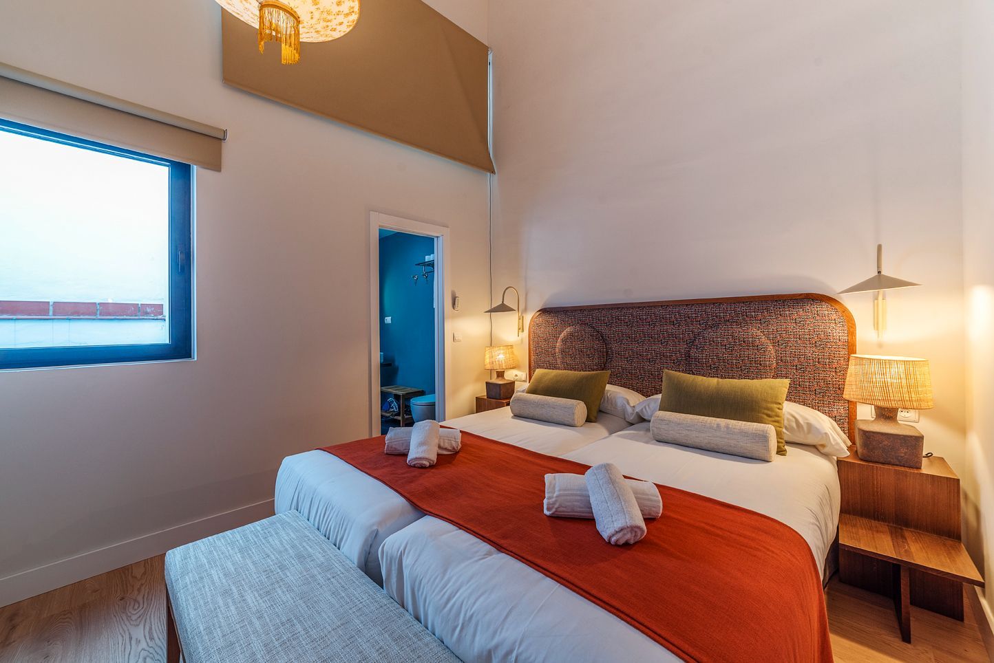 Habitación acogedora con cama, manta naranja, puerta azul y luz en la ventana.