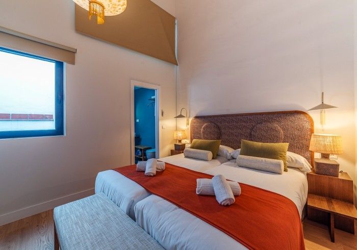Dormitorio moderno con una cama grande, manta naranja, lámparas de noche y puerta azul en una habitación luminosa.