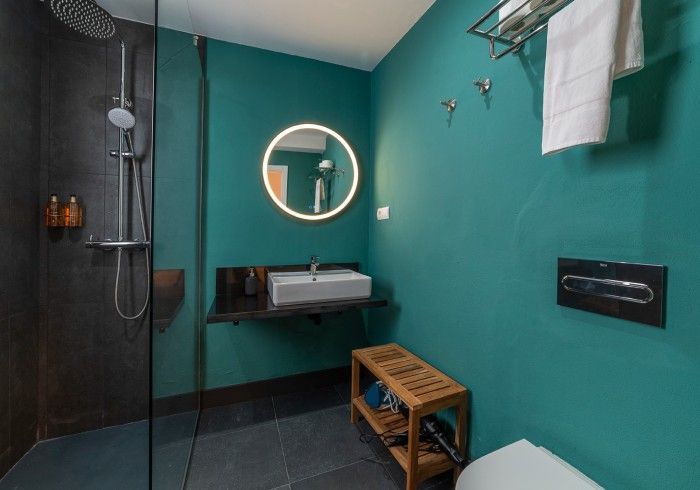 Baño moderno con paredes color turquesa, ducha negra, espejo de pared, lavabo, toallero y banco con taburete de madera.