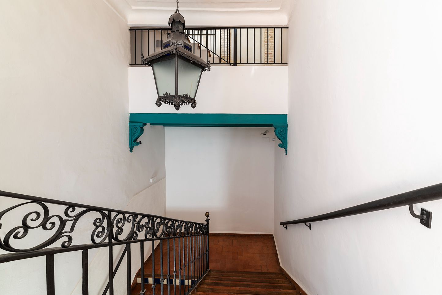 Escalera con barandilla de hierro forjado, escalones de madera y una linterna colgante bajo un arco de color verde azulado.