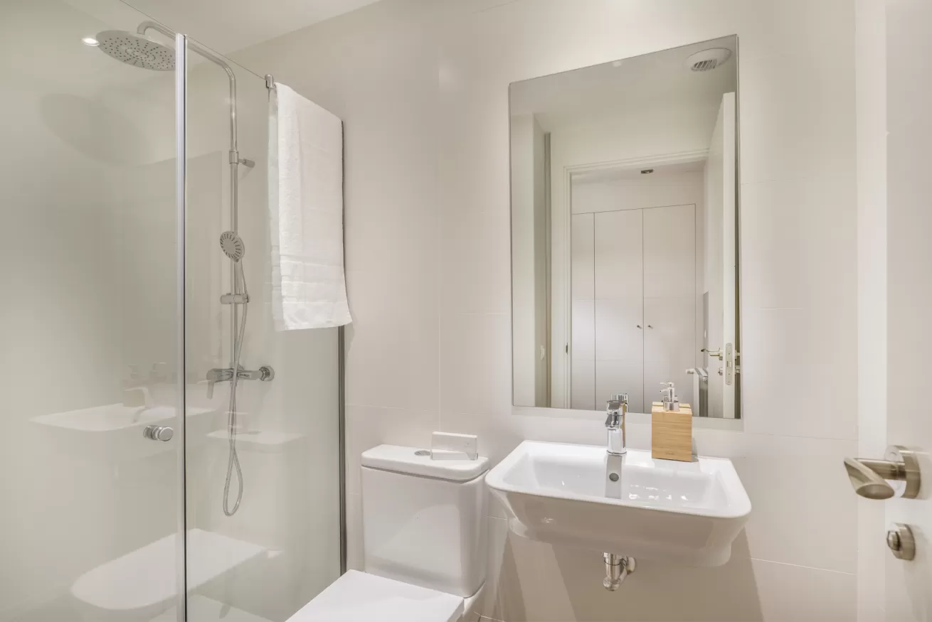 Baño de un blanco brillante con ducha, inodoro y lavabo bajo un gran espejo.