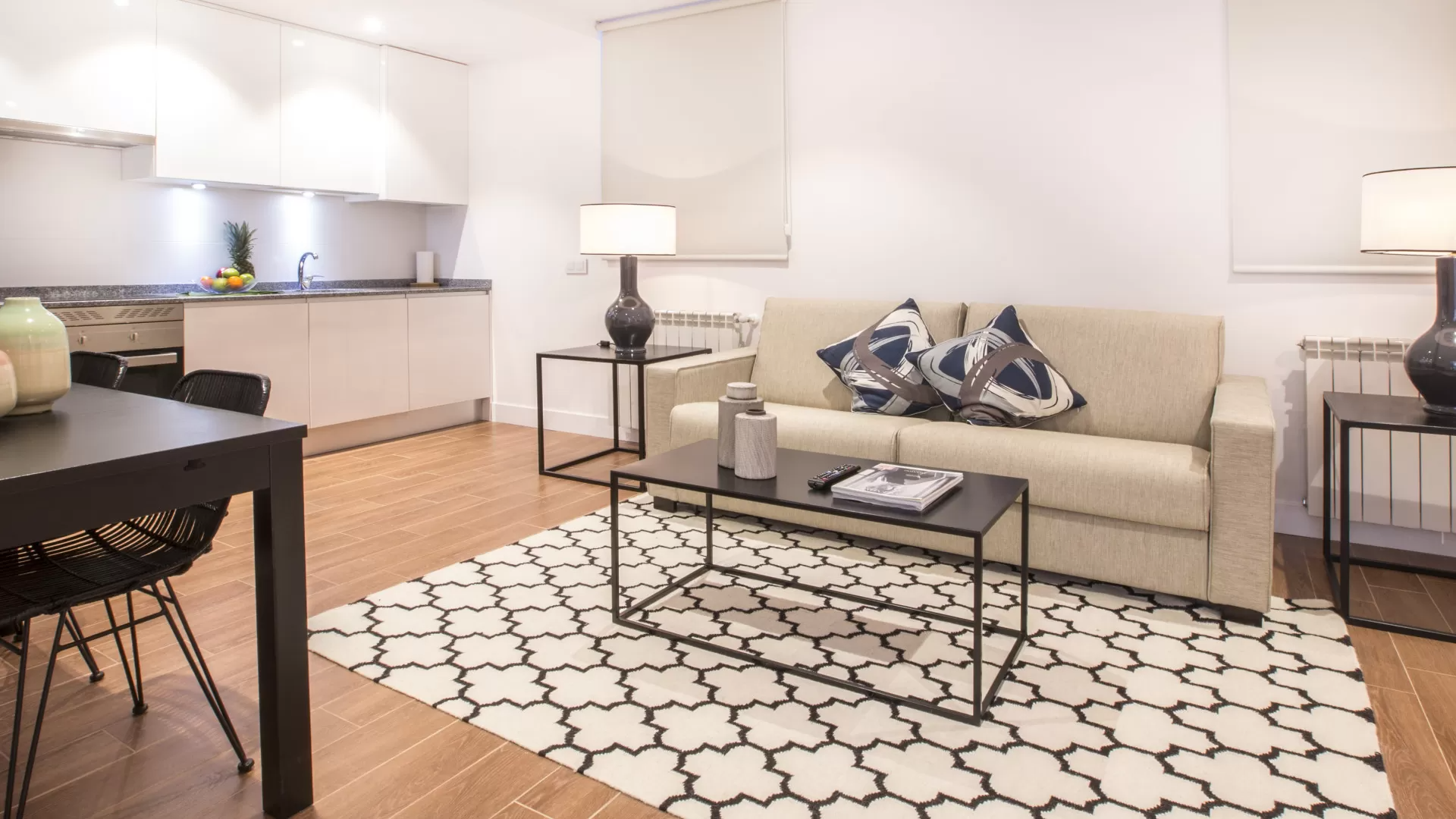 Sala de estar moderna de planta abierta con sofá beige, alfombra estampada, mesa de centro y cocina al fondo.