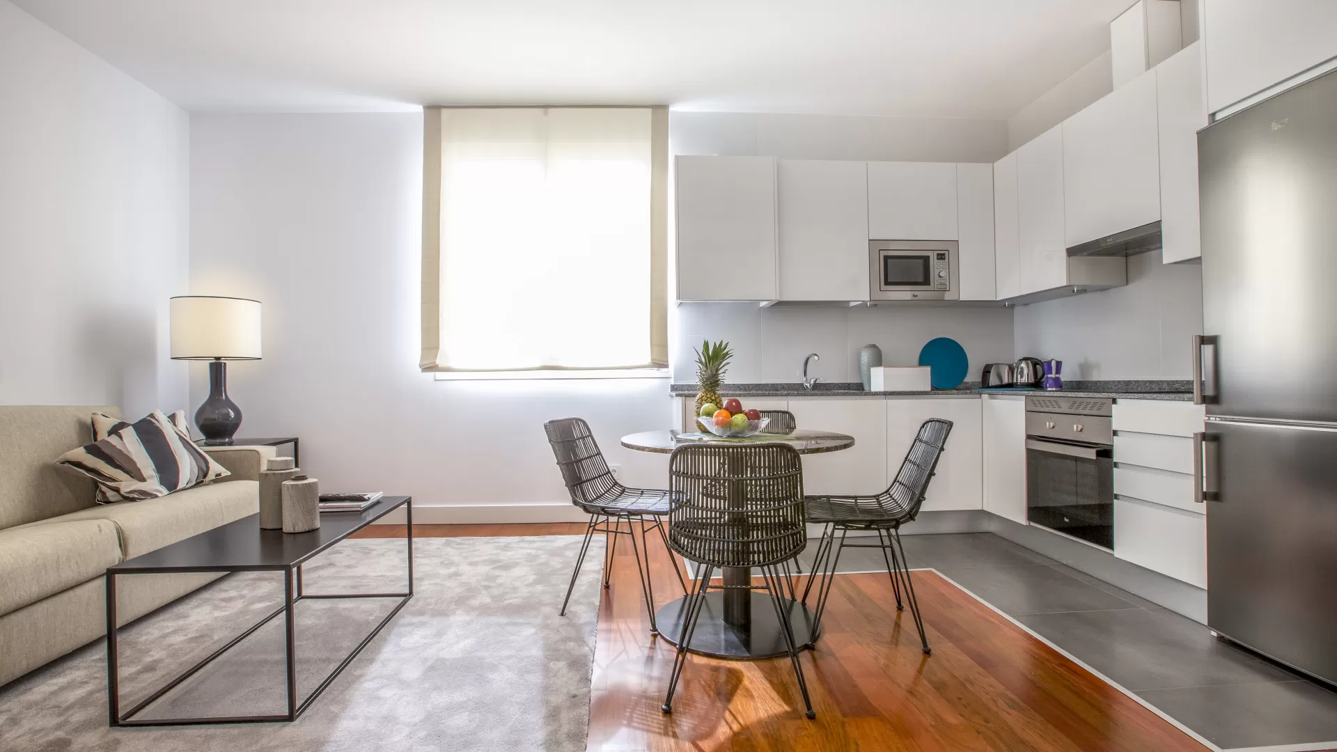 Estudio luminoso y moderno con sofá gris, mesa de centro de cristal, juego de comedor y armarios de cocina blancos.