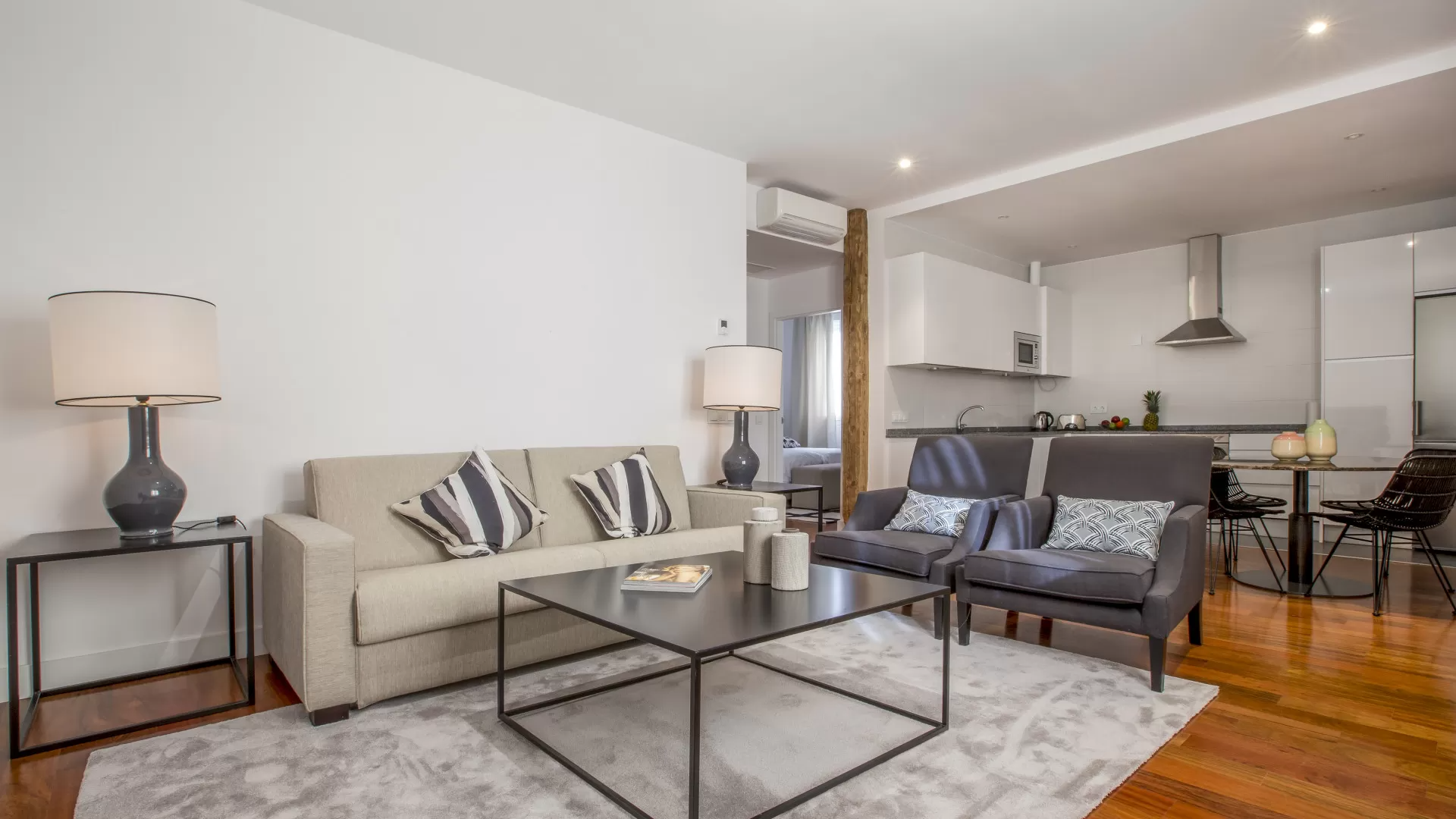 Sala de estar moderna con sofá beige, sillones oscuros, mesa de centro de cristal y cocina abierta al fondo.