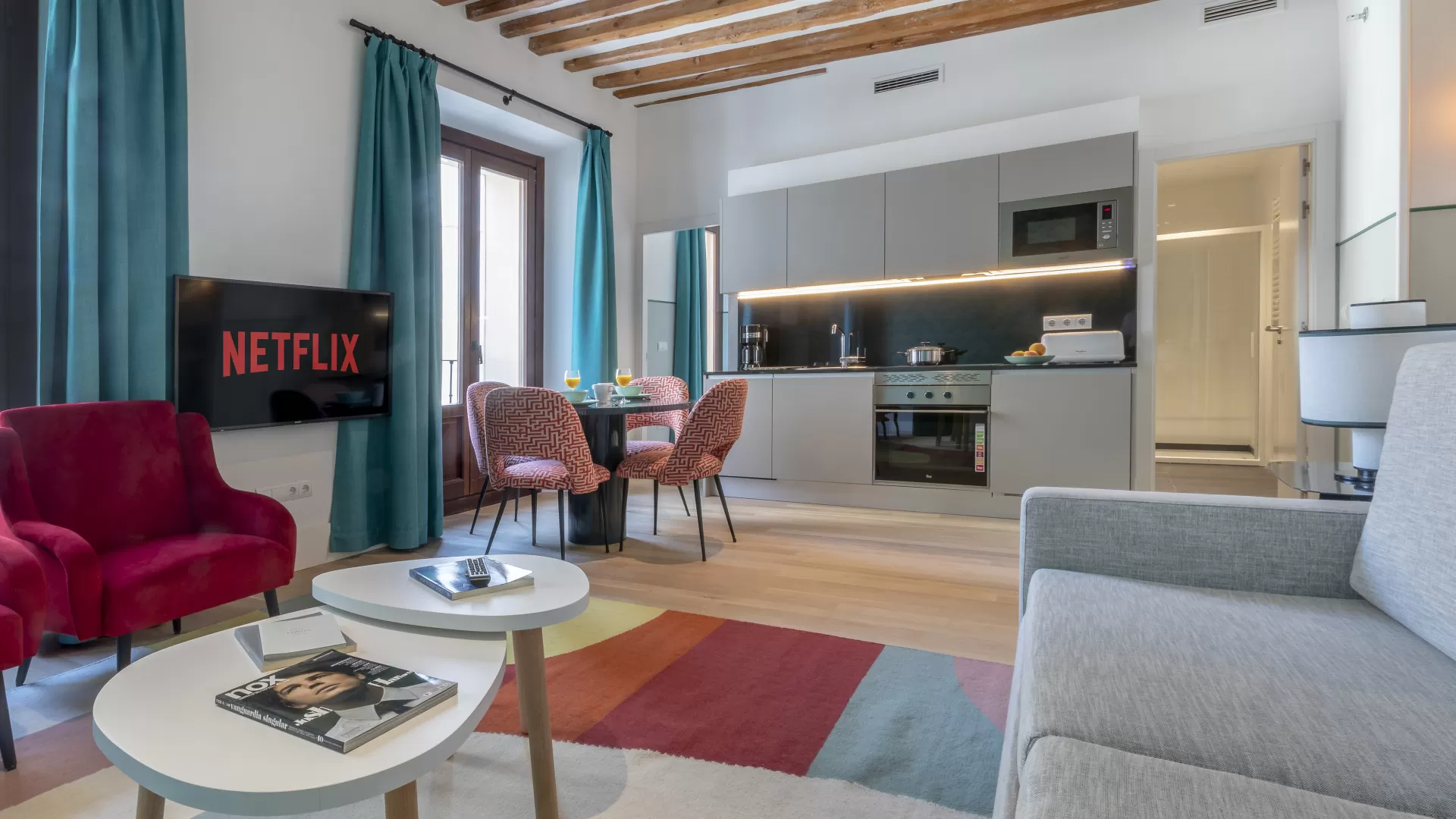 Luminoso salón moderno con sofá, sillones, mesa de centro, cocina y televisión con Netflix.