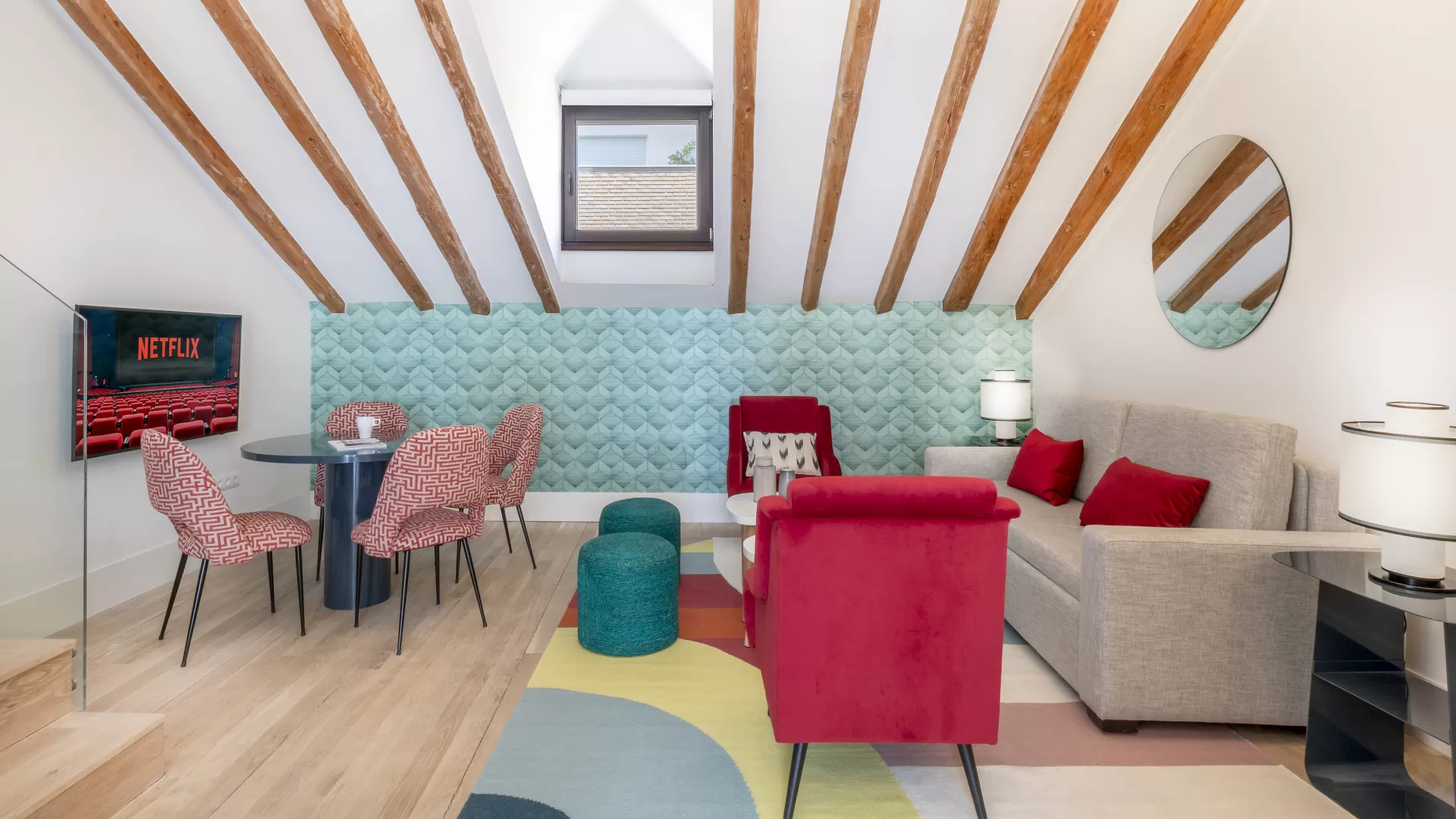 Sala de estar moderna y luminosa con banco color turquesa, sillas rojas, sofá beige y vigas de madera en el techo.