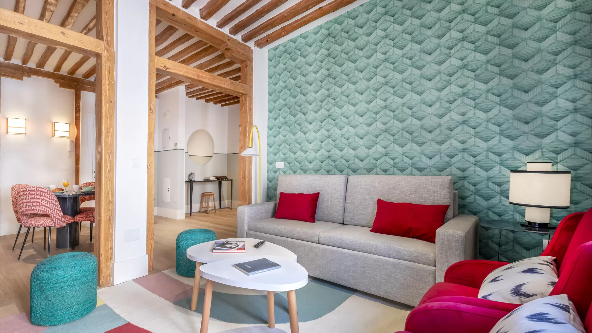 Luminoso salón con pared estampada en color turquesa, sofá gris, cojines rojos, mesa redonda y vigas de madera.