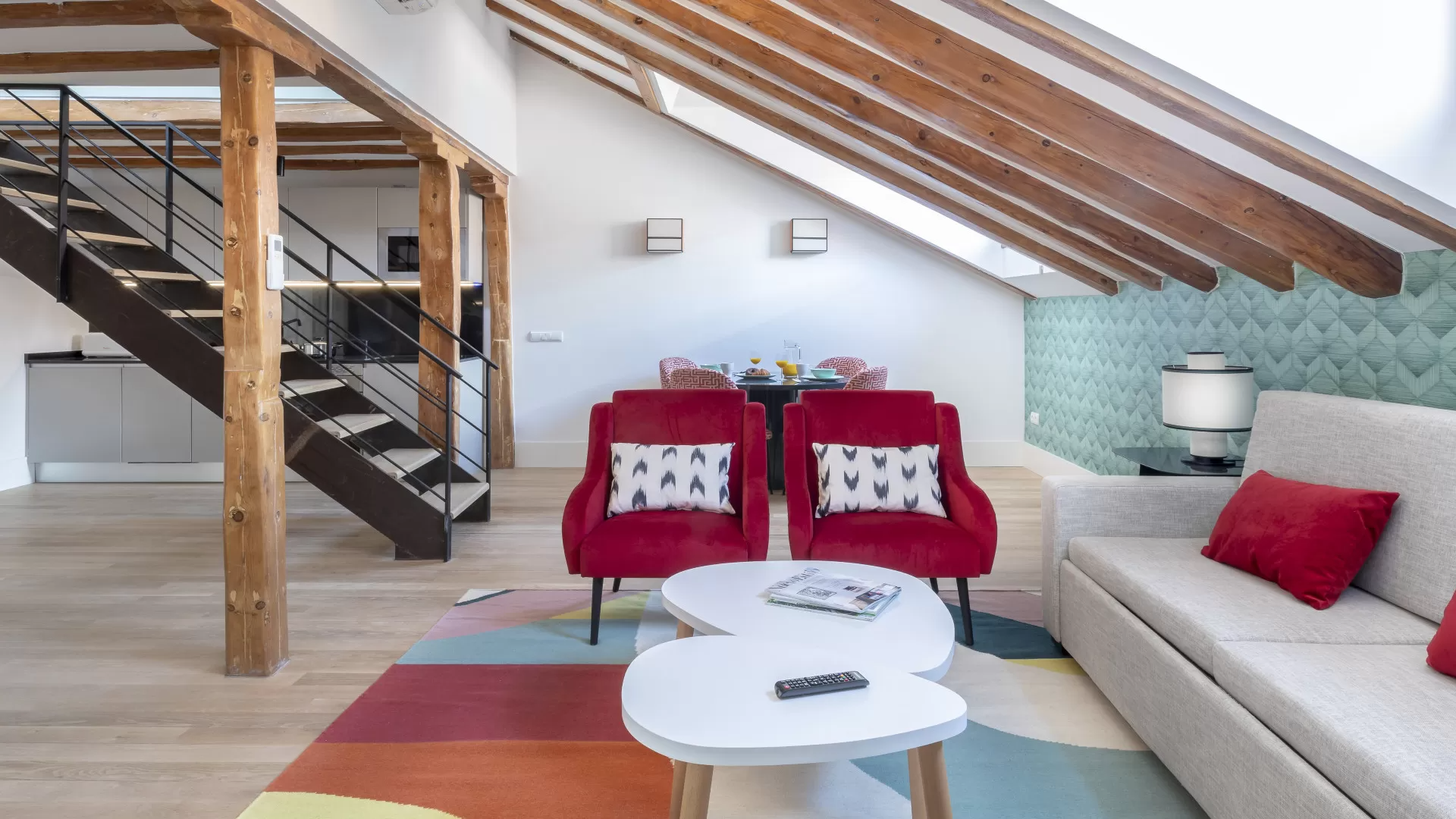 Sala de estar tipo loft moderna con vigas a la vista, escalera, sofá beige, sillas rojas y alfombra colorida.