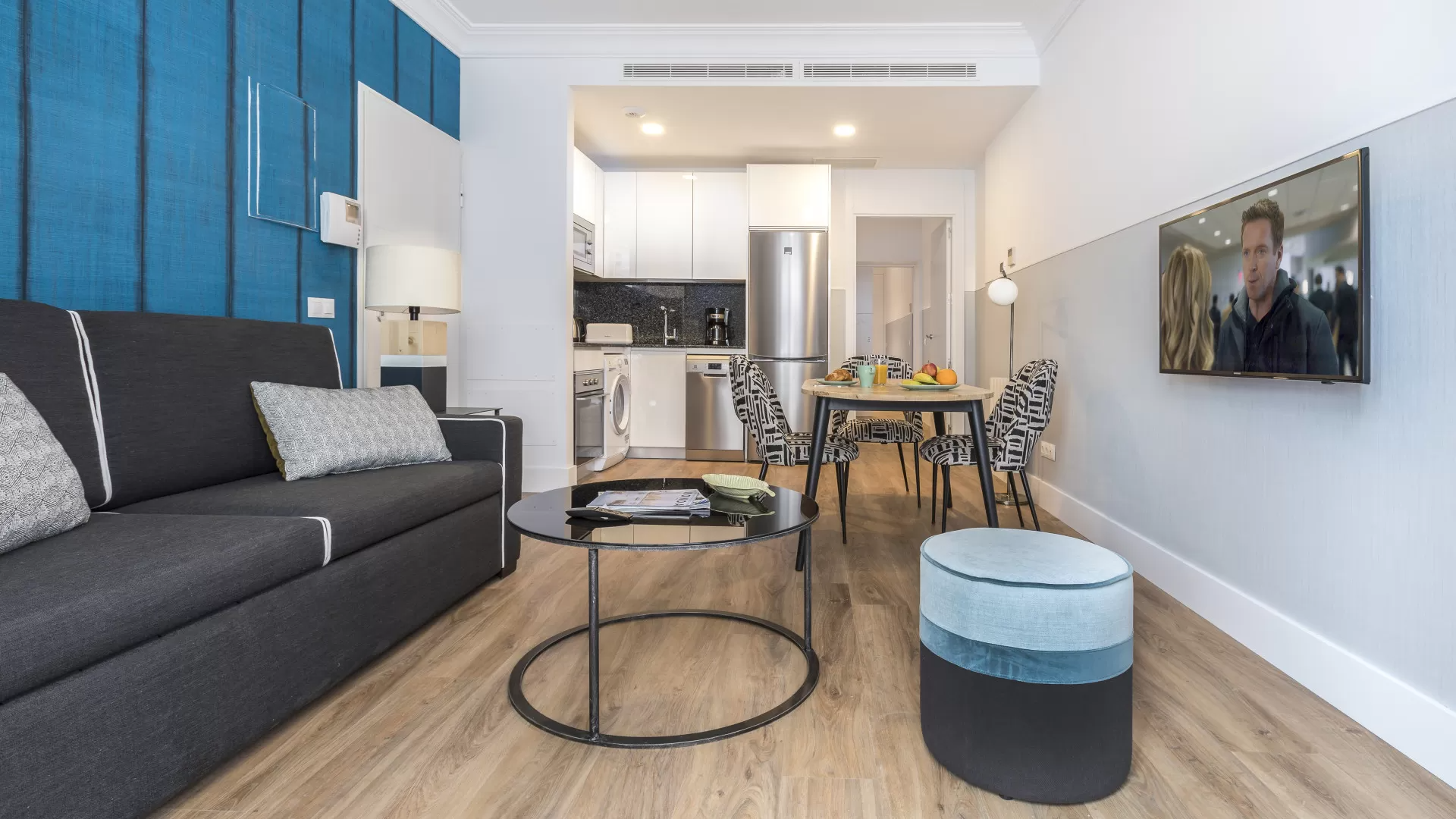 Sala de estar moderna con sofá gris, pared decorativa azul, mesa de centro redonda y televisor montado en la pared.