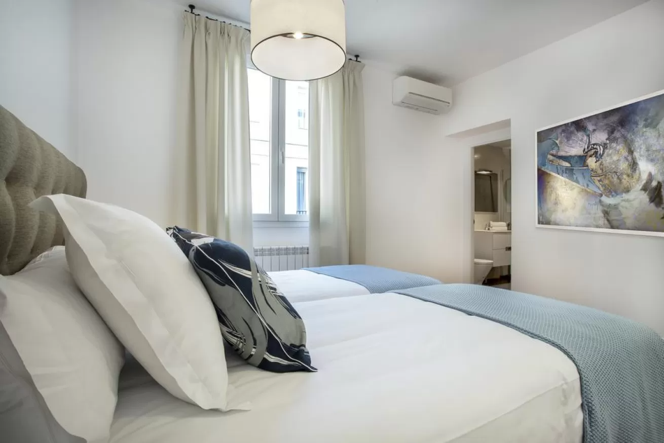 Habitación luminosa con cama blanca, manta azul, ventana, cuadro en la pared y aire acondicionado.