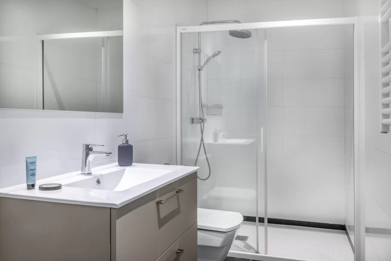 Baño moderno blanco con lavabo, espejo, inodoro y cabina de ducha de cristal.