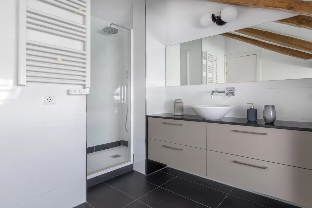Baño moderno con ducha a ras de suelo, espejo grande, tocador beige y suelo de baldosas negras.