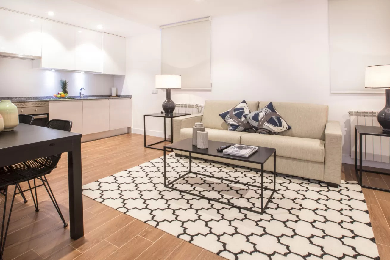 Salón moderno con sofá beige, alfombra estampada, mesa de centro de cristal, lámparas de pie y cocina abierta.