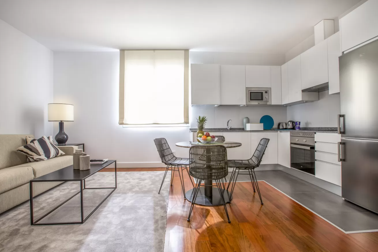 Luminoso salón-cocina moderno de planta abierta con sofá gris, mesa de comedor, armarios blancos y suelo de madera.