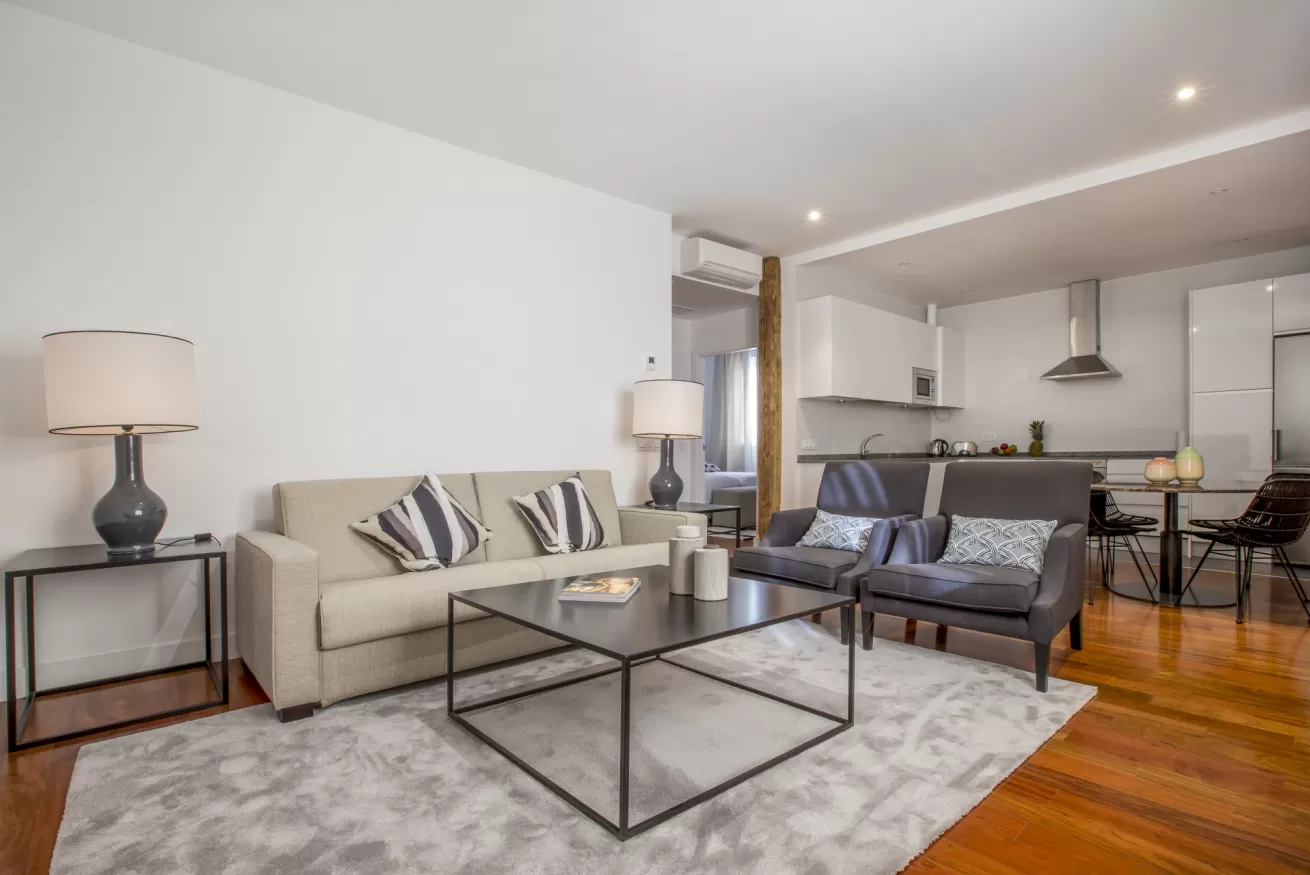 Sala de estar moderna de planta abierta con sofá beige, sofá de dos plazas gris, mesa de centro de cristal y cocina al fondo.
