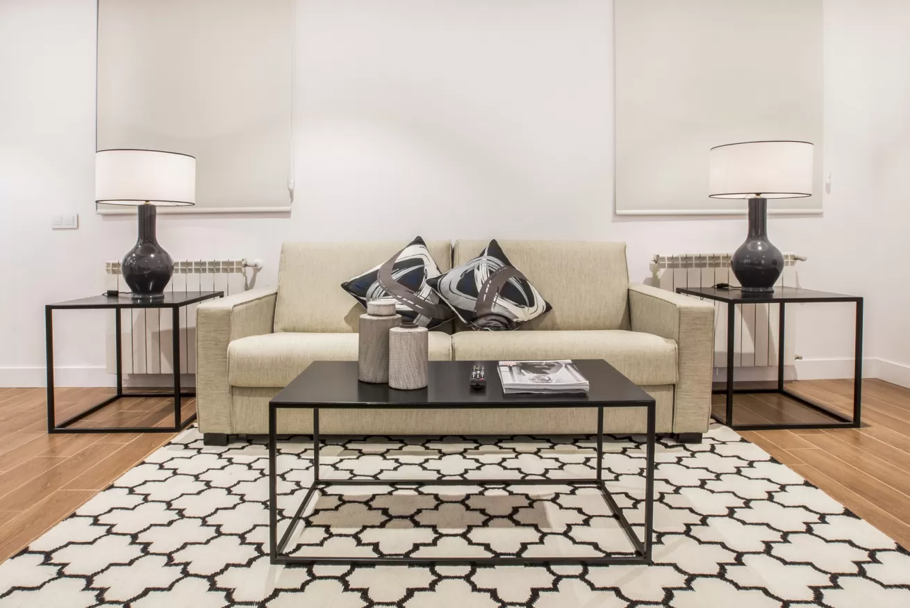 Sala de estar moderna con sofá beige, alfombra estampada y mesitas auxiliares negras con lámparas.