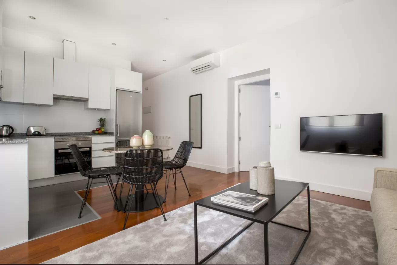 Salón moderno de planta abierta con cocina, mesa de comedor, televisor montado en la pared y zona de sofá gris.