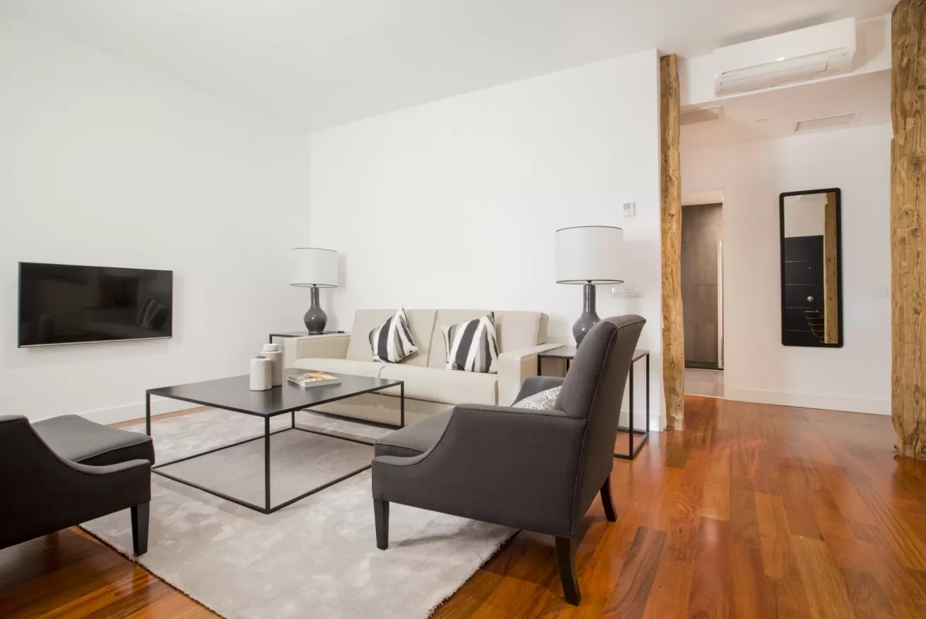 Sala de estar moderna con sofá blanco, dos sillones oscuros, mesa de centro de cristal, televisor de pared y suelo de madera.