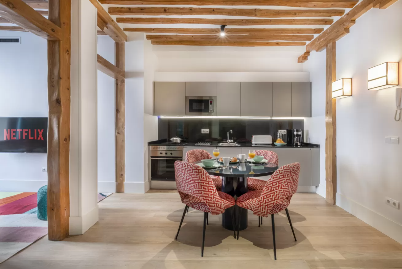 Cocina-comedor moderna con vigas a la vista, mesa redonda, sillas rojas y armarios oscuros.