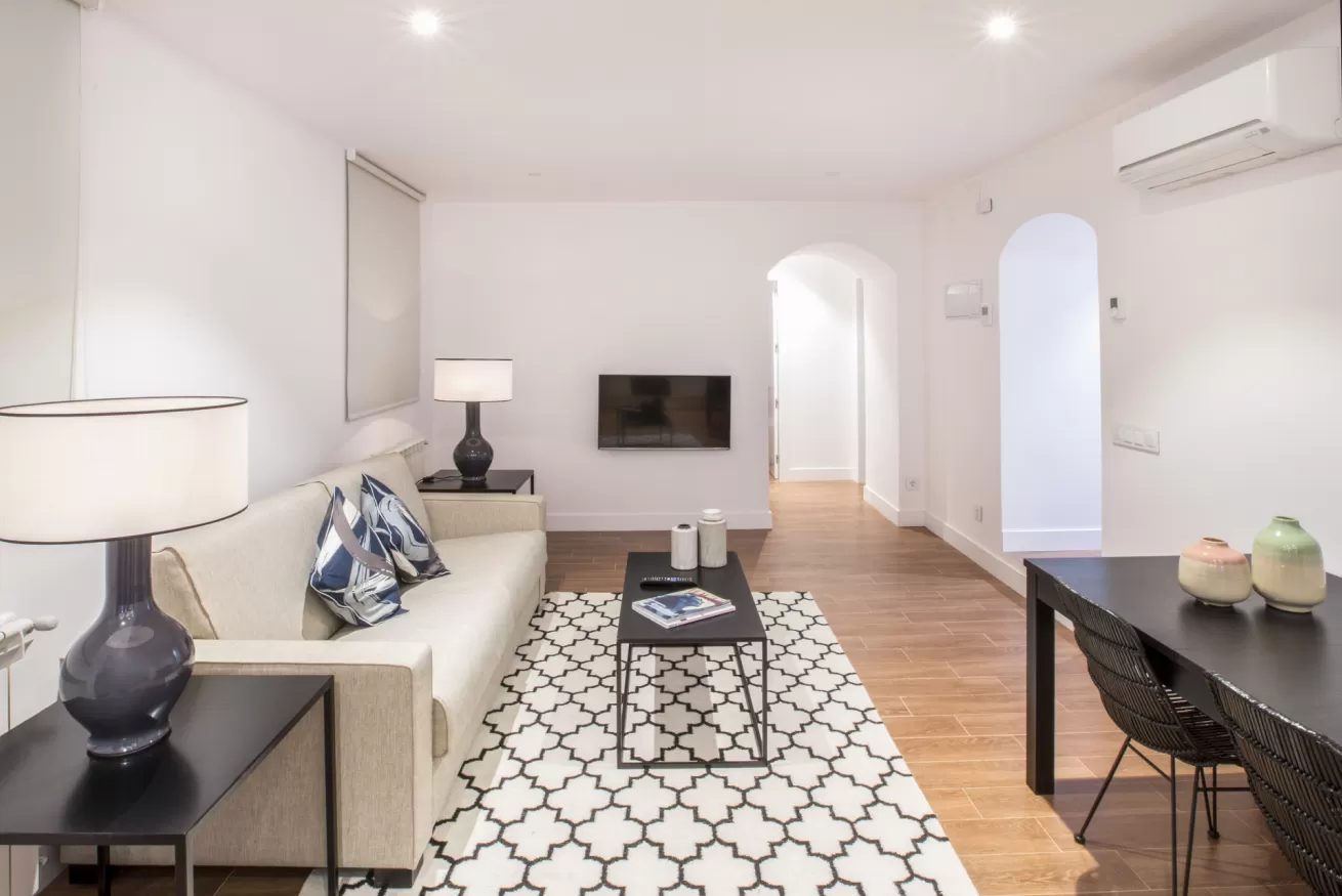 Luminoso salón con sofá beige, mesa de centro negra, alfombra estampada, televisor montado en la pared y mesa.