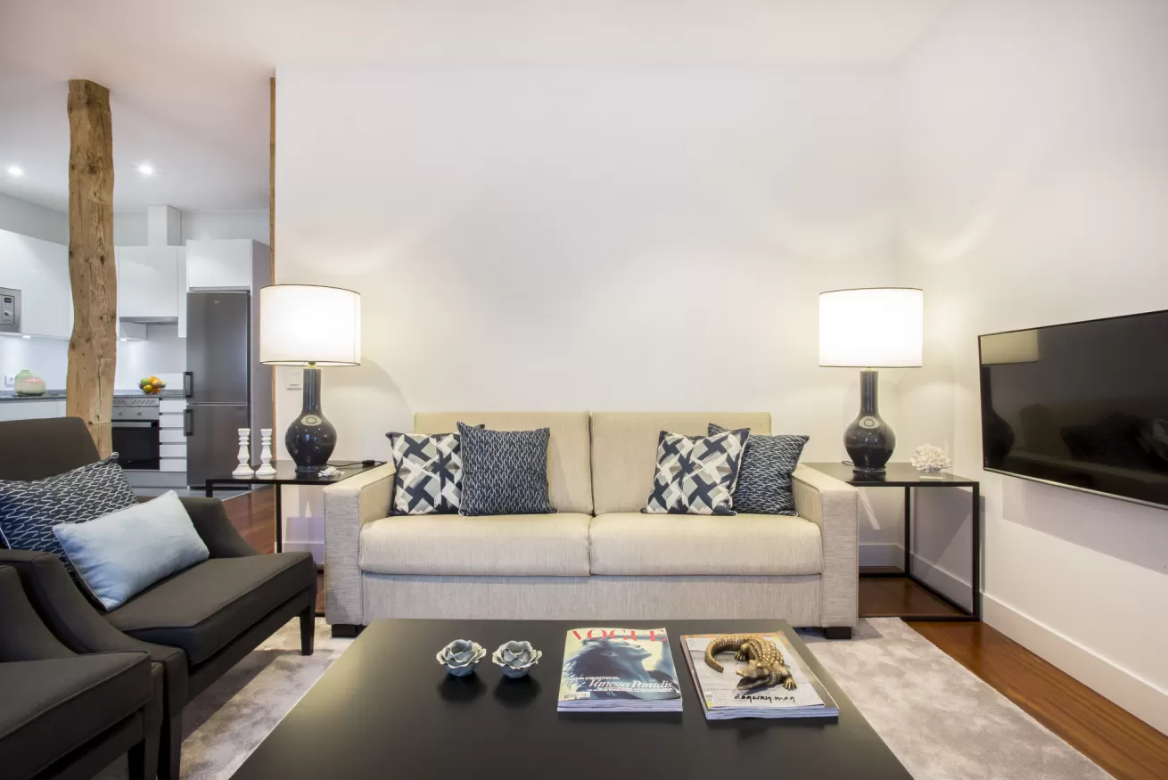 Sala de estar moderna con sofá beige, mesa de centro negra, televisor montado en la pared y dos lámparas.