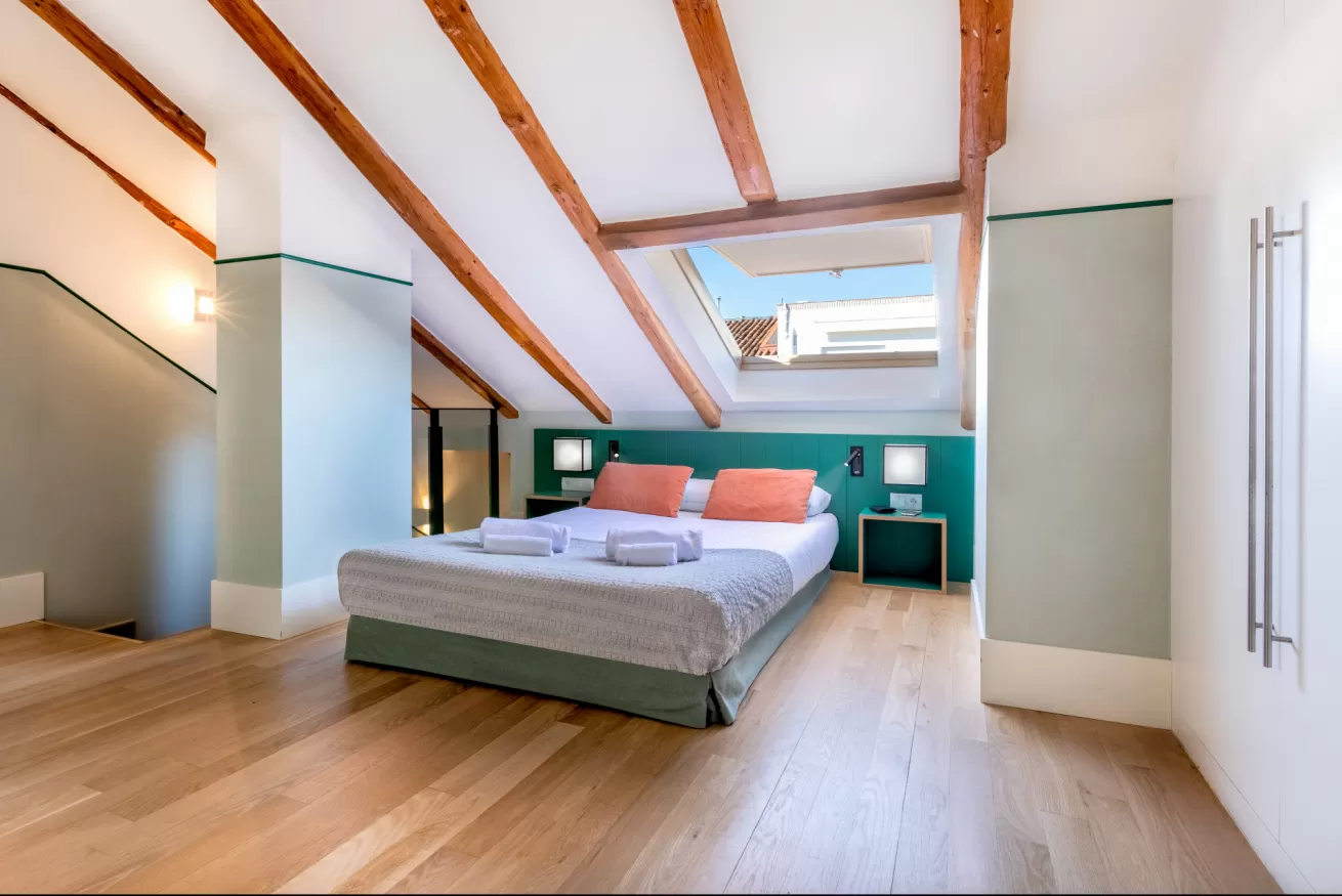 Luminoso dormitorio abuhardillado con una cama grande, pared decorativa de color turquesa, tragaluz y vigas de madera.