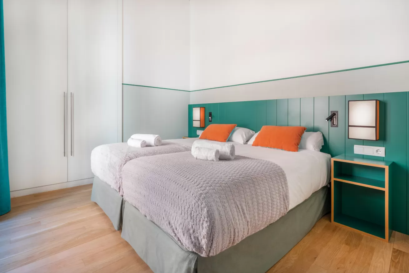 Dormitorio moderno con una cama grande, ropa de cama gris, almohadas naranjas y una pared decorativa de color verde azulado.
