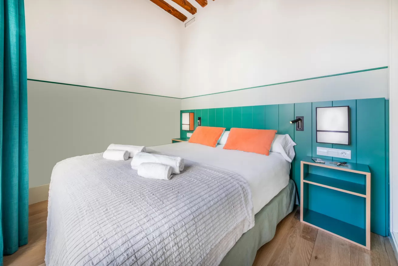 Habitación luminosa con una cama grande blanca, almohadas color coral, paredes turquesa y una mesita de noche azul.