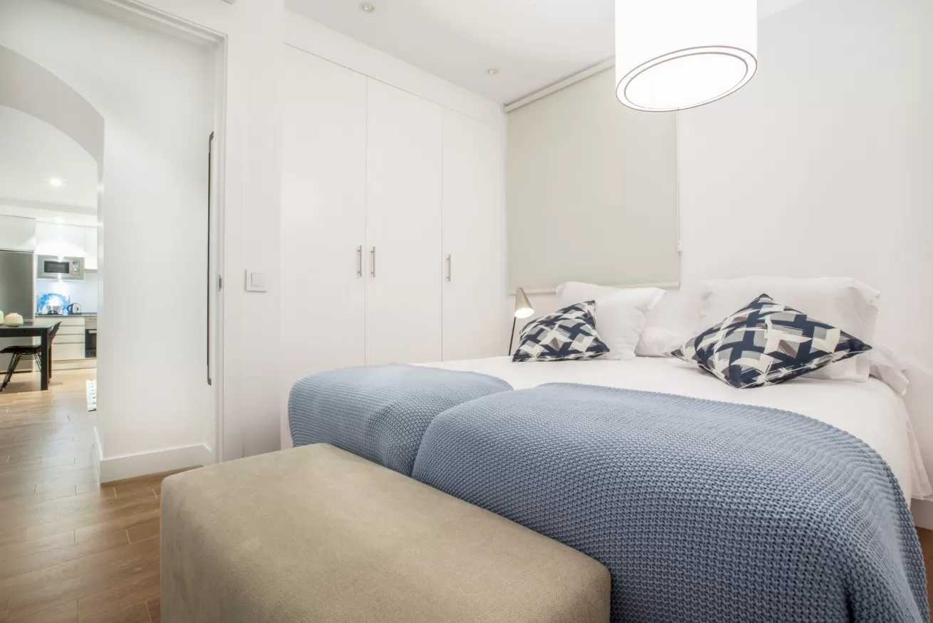 Habitación blanca y luminosa con una cama bien hecha, una colcha azul y un banco beige a los pies.