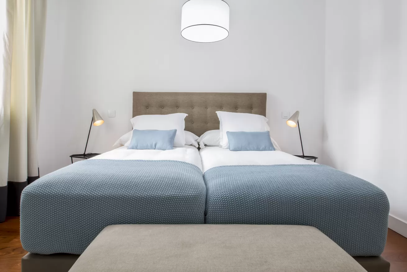 Dormitorio minimalista con una cama azul, almohadas blancas, lámparas de noche y un banco beige a los pies de la cama.