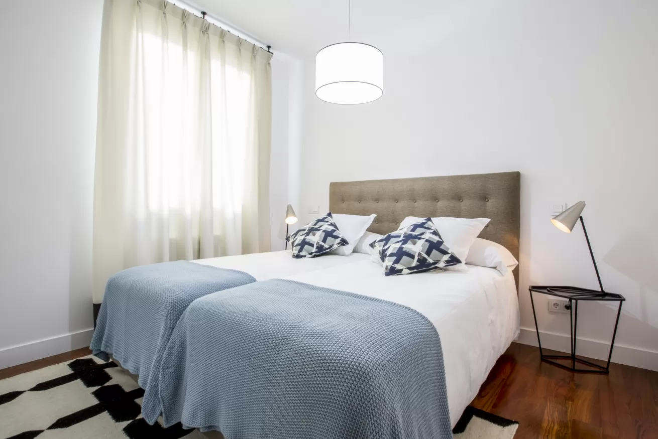 Habitación luminosa con dos camas, edredones azules, cortinas blancas y una lámpara de noche.
