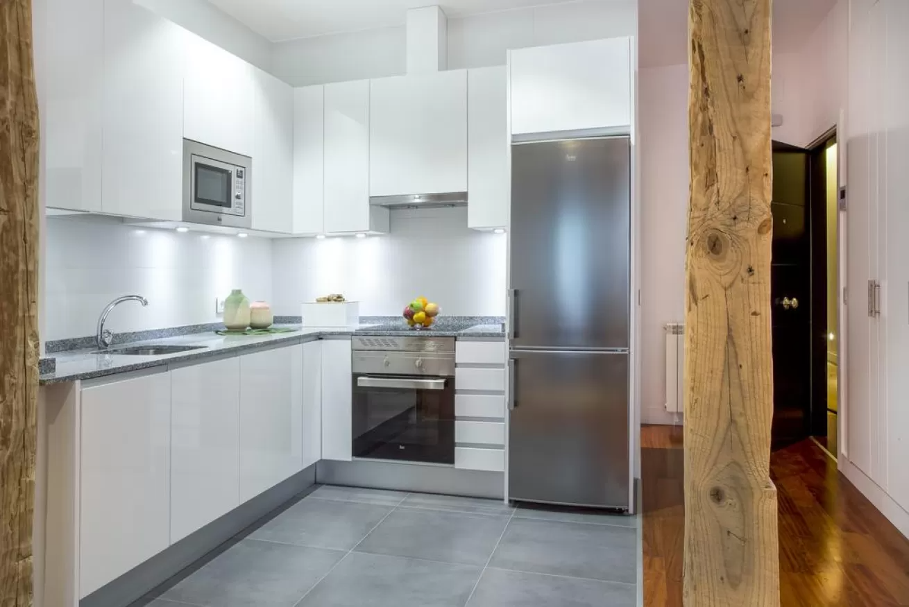 Cocina moderna blanca con electrodomésticos de acero inoxidable, baldosas grises en el suelo y una puerta con viga de madera.