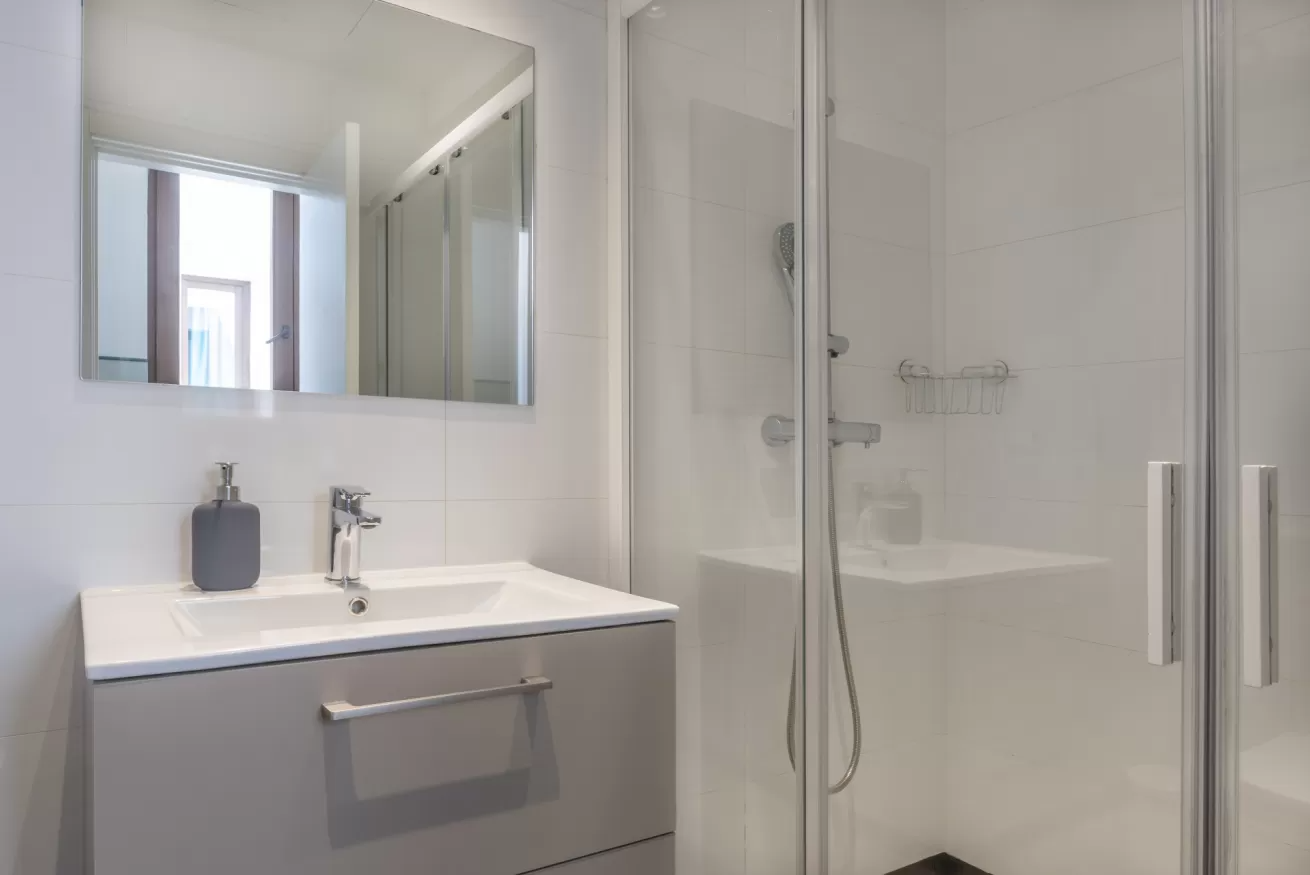 Baño moderno blanco con lavabo, espejo, cabina de ducha y puerta de cristal.