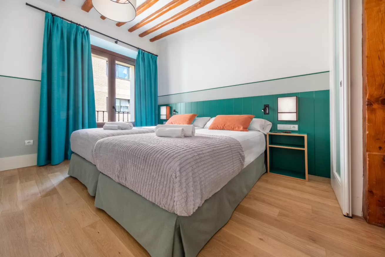 Habitación luminosa con cama doble, cortinas color turquesa, suelo de madera y una pared decorativa turquesa con espejo.
