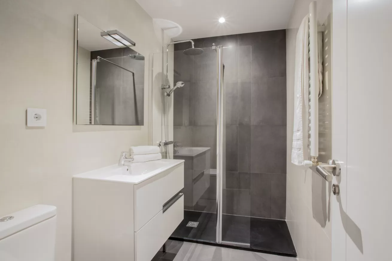 Baño moderno con tocador blanco, espejo, inodoro y ducha con mampara de cristal y azulejos oscuros.