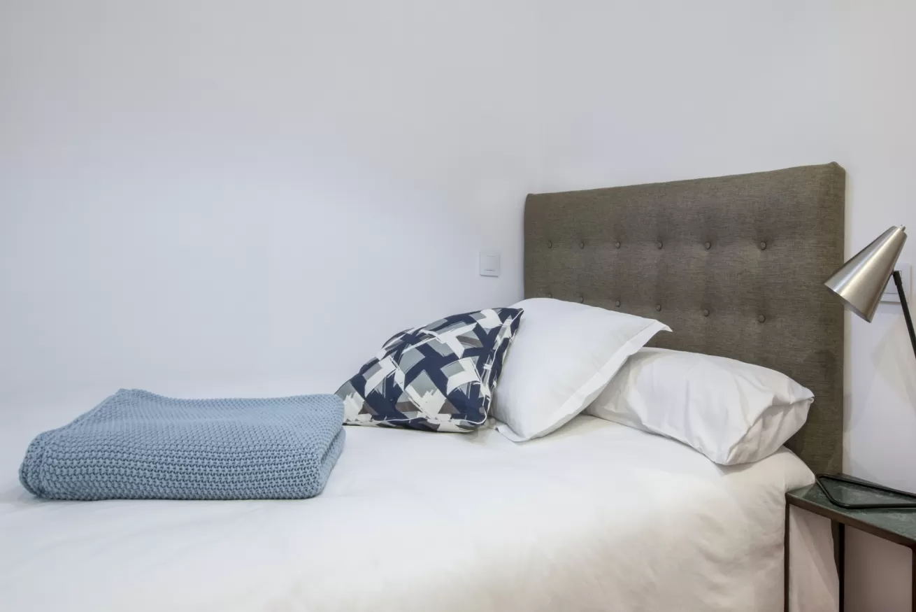 Cama bien hecha con sábanas blancas, manta y almohadas azules, junto a un cabecero acolchado gris y una lámpara de noche.