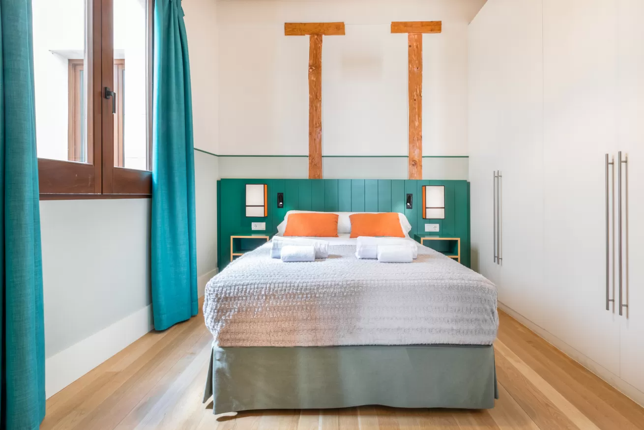 Habitación luminosa con cama, cabecero turquesa, detalles en naranja y suelo de madera junto a una ventana.