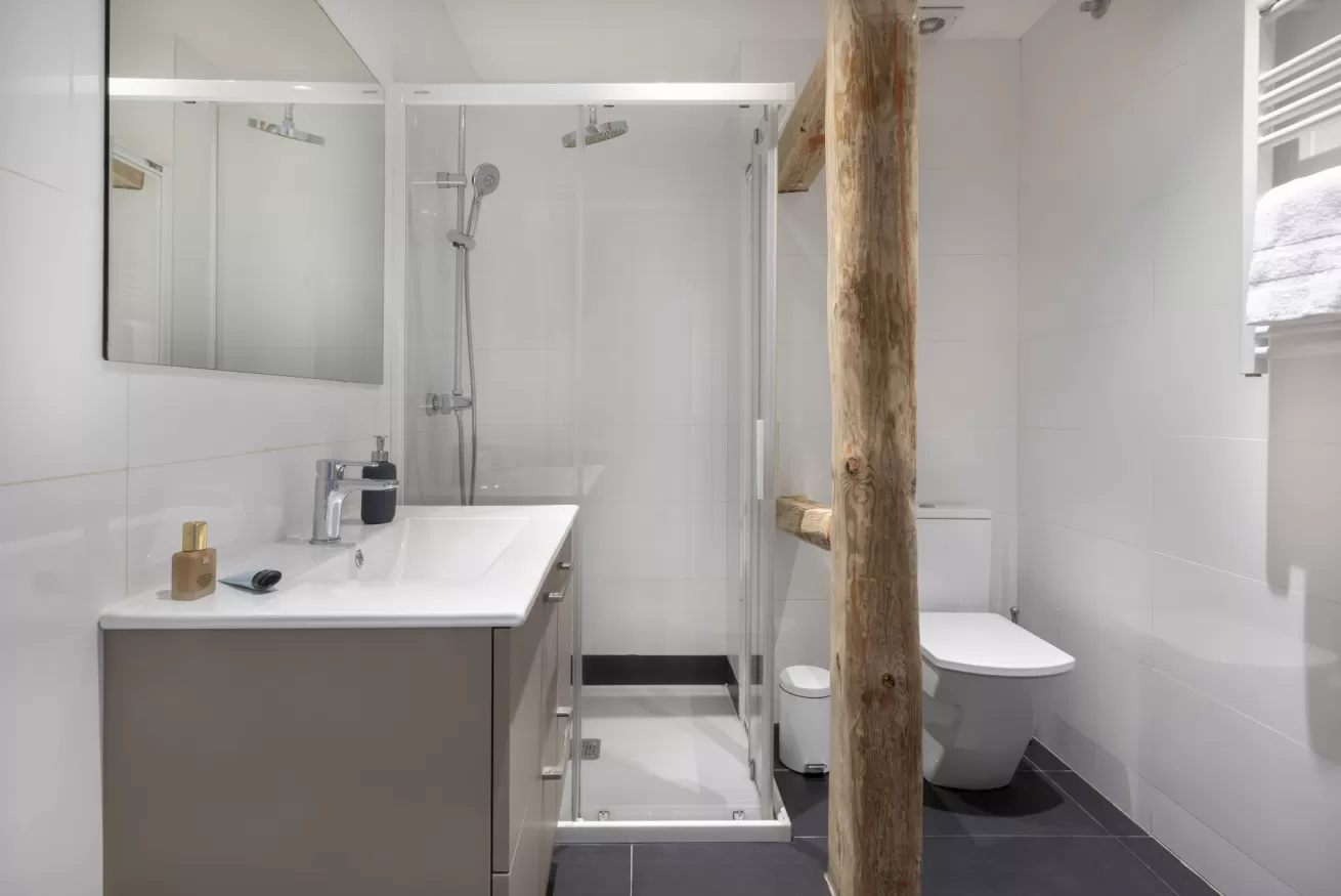 Baño moderno con azulejos blancos, ducha a ras de suelo, lavabo con mueble, inodoro y viga de soporte de madera.