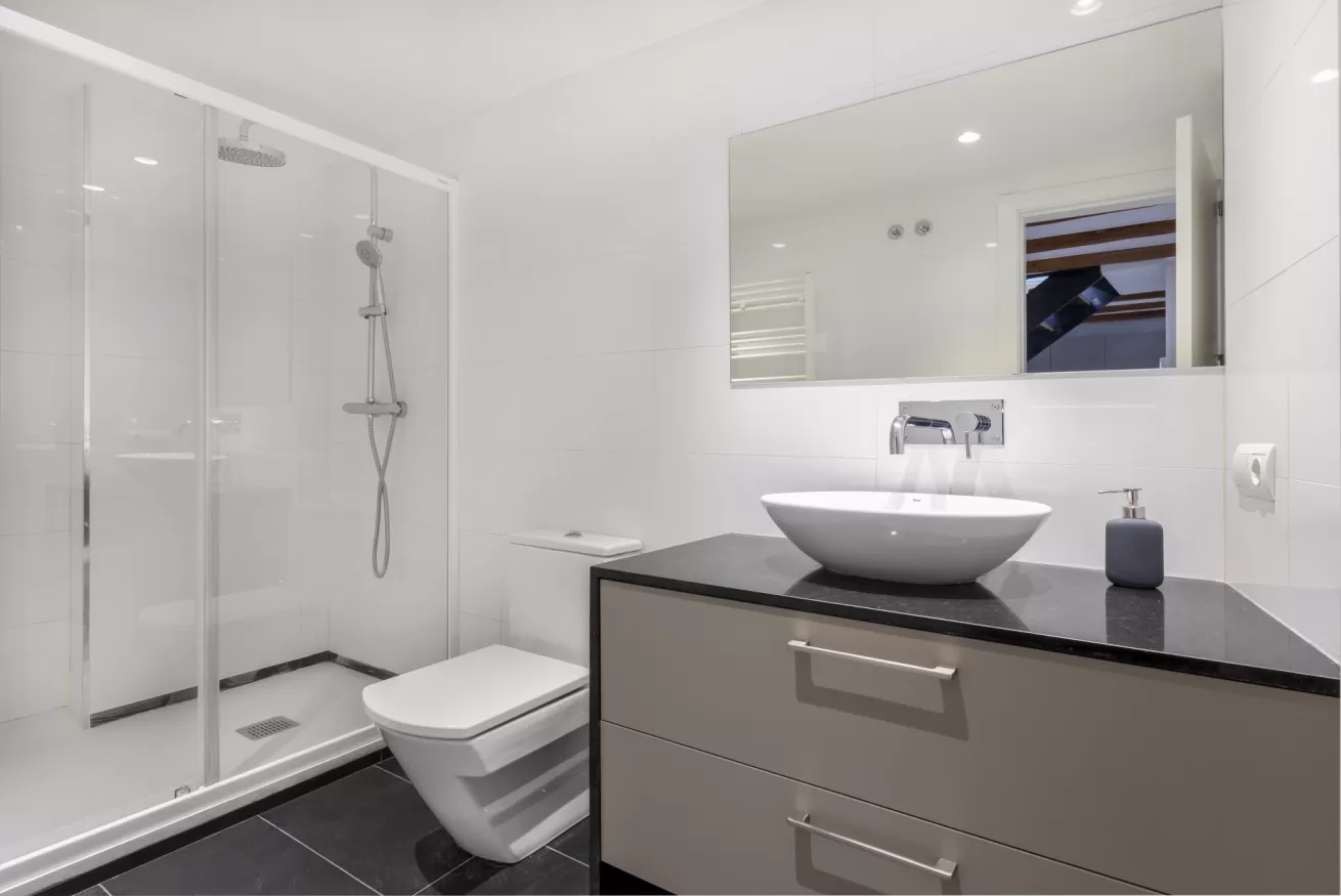 Baño moderno de color blanco con ducha de cristal, inodoro suspendido y tocador negro con lavabo sobre encimera.