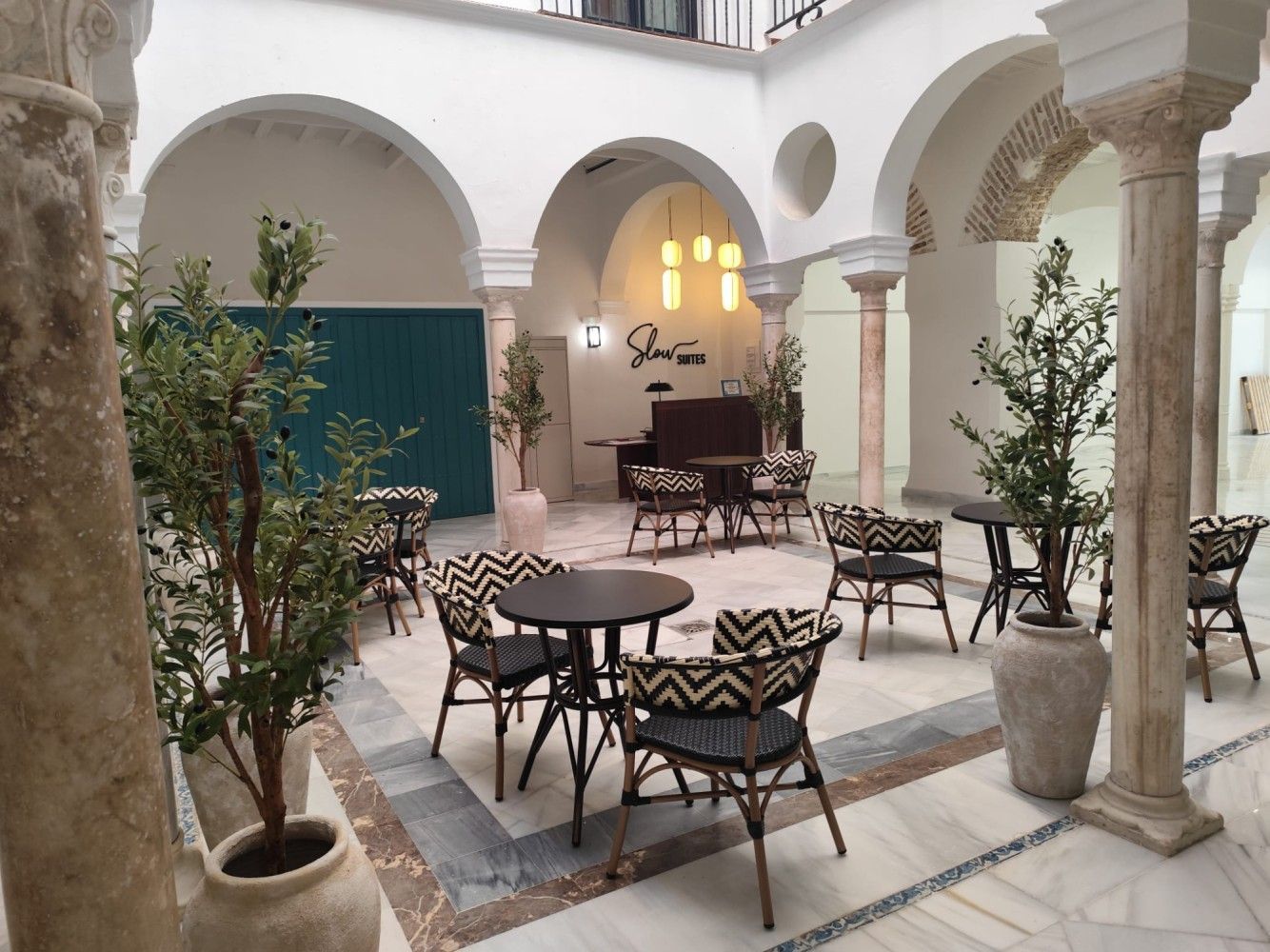 Cafetería soleada con patio, paredes blancas arqueadas, mesas y sillas de metal negro, plantas en macetas.