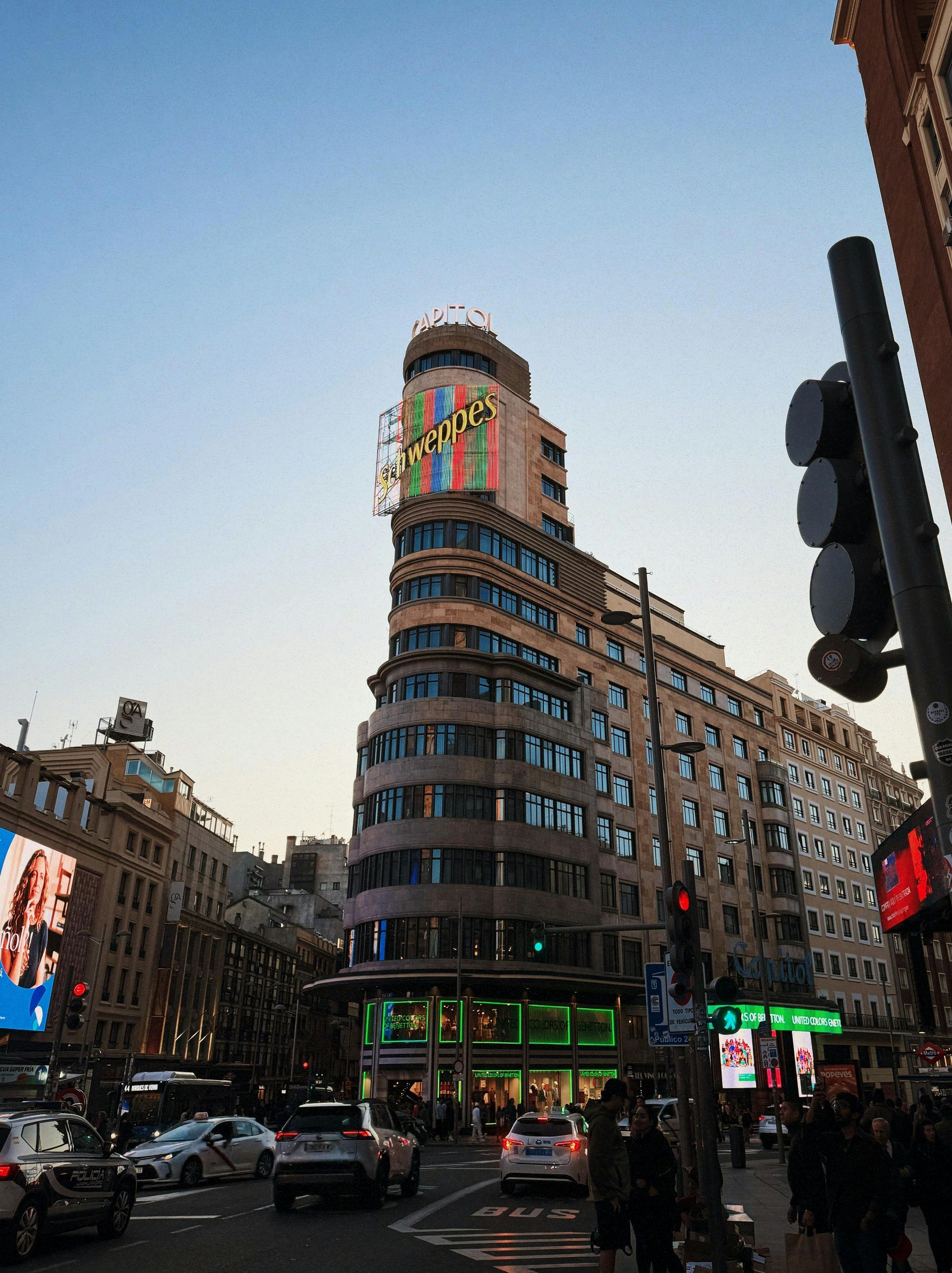 Edificio curvo de la ciudad con luces de colores en el tejado en una calle concurrida al atardecer bajo un cielo despejado.