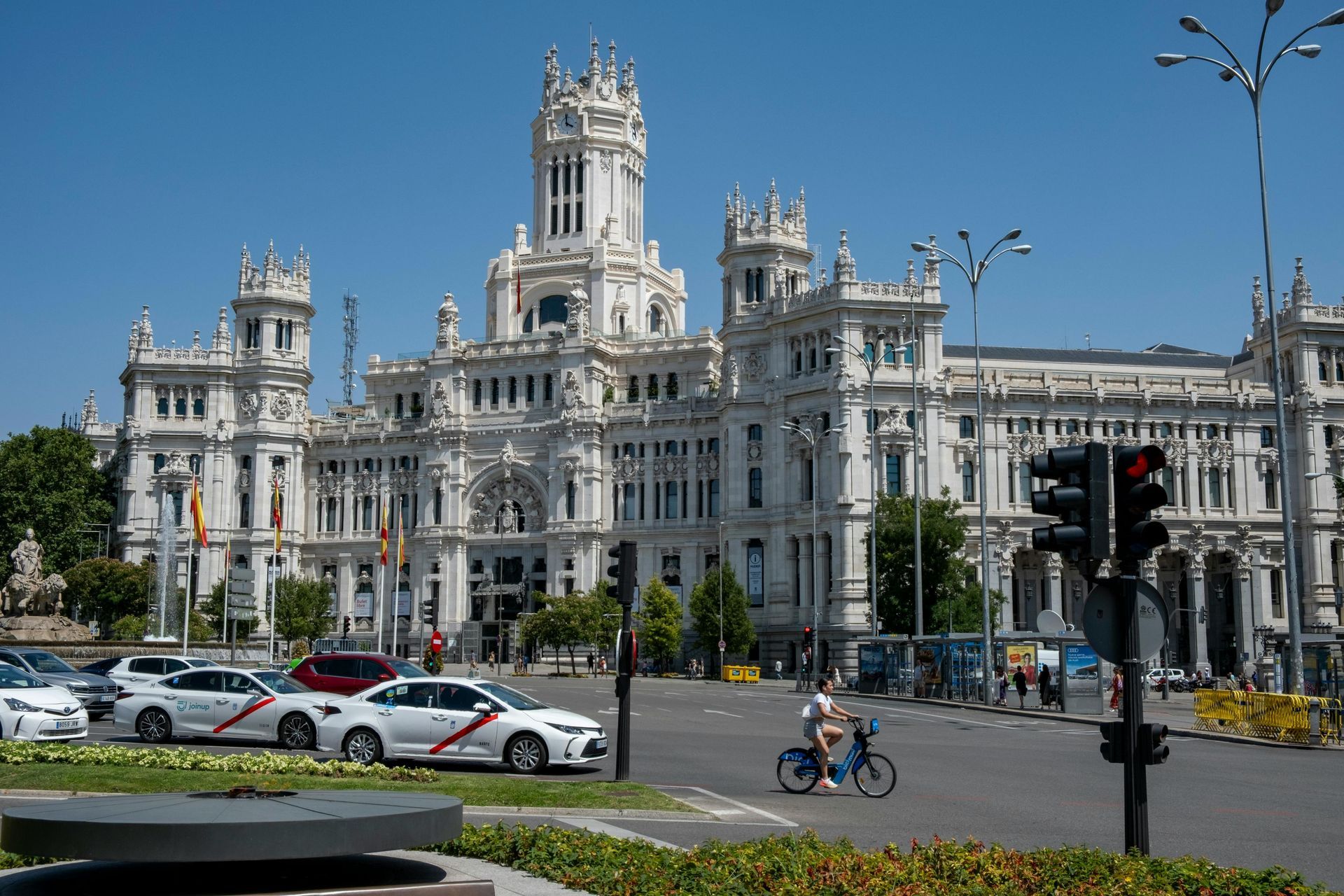 Edificio cívico blanco y ornamentado con torres, coches y un ciclista en una soleada calle de la ciudad.