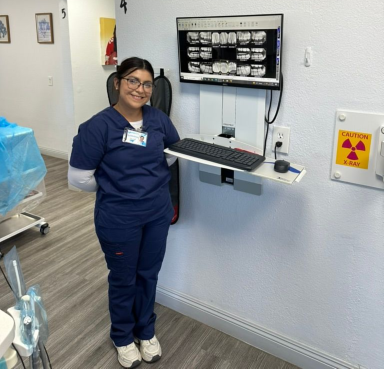 Dental assistant smiles next to a monitor showing dental x-rays in a dentist's office.
