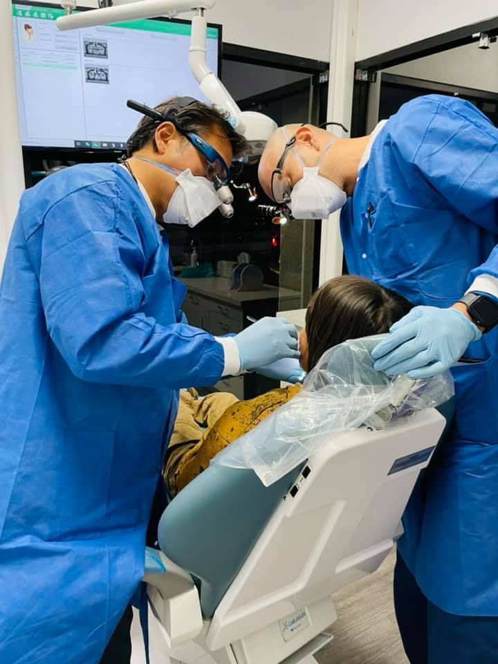 Two dentists in blue scrubs examining a patient in a dental chair.