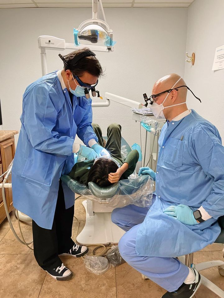 Two dentists in blue scrubs examine a patient in a dental chair.